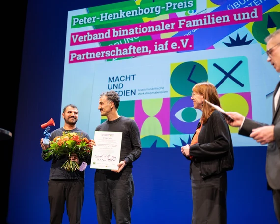 Bühnenaufnahme der Preisverleihung: Zwei Personen halten Blumenstrauß, Urkunde und rote Megafon-Statuette, daneben weitere Personen. Im Hintergrund Leinwand mit Schrift „Peter-Henkenborg-Preis – Verband binationaler Familien und Partnerschaften, iaf e.V.“