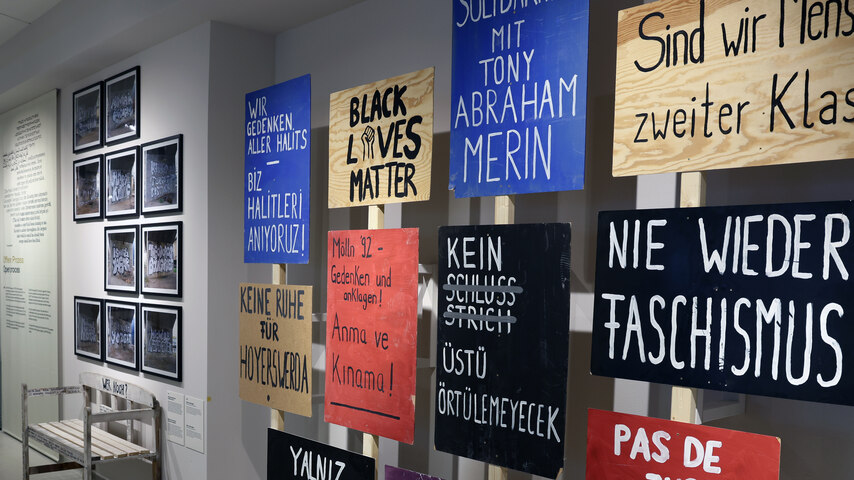 Mehrere Schilder mit Botschaften sind in der lrechten Bildhälfte zu sehen. In der linken Bildhälfte ist eine Wand mit Bilderrahmen.