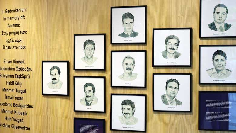 Zu sehen sind die gezeichneten Porträts der Opfer des NSU in schwarz-weiß auf holzfarbenen Untergrund.