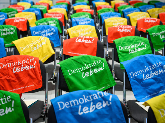 Eine Vielzahl von Stühlen über den Rückenlehnen Baumwolltaschen in verschiedenen Farben liegen. Der Schriftzug Demokratie leben ist auf diesen abgedruckt.