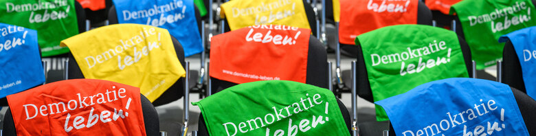 Auf den Bild sind Stühle zu sehen. Über die Rückenlehnen sind Baumwollbeutel in verschiedenen Farben gelegt. Auf ihnen ist der Schriftzug Demokratie leben zu lesen.