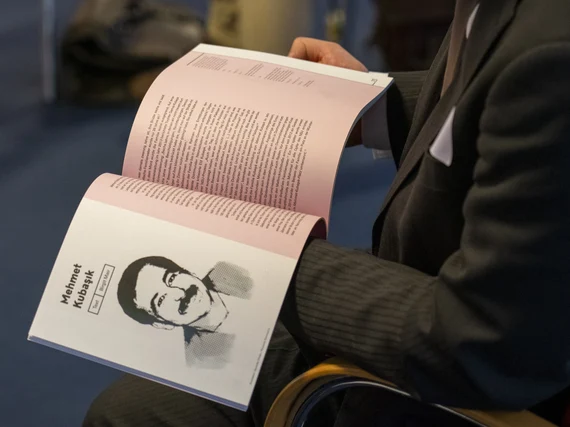 Person im Anzug sitzt auf einem Stuhl und hält eine geöffnete Broschüre mit rosa Seiten auf dem Schoß; sichtbar ist eine Textseite und eine Seite mit Porträtzeichnung und Namen.