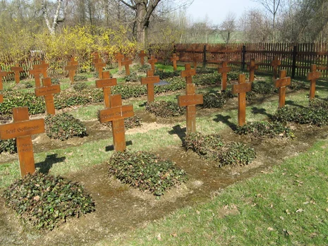 Feld mit Reihen kleiner Kriegsgräber: Auf jedem Grab steht ein rostbraunes Metallkreuz, die Gräber sind mit Bodendeckern bepflanzt, im Hintergrund Bäume, Sträucher und ein Holzzaun.