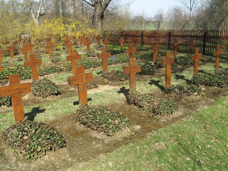 Das Bild zeigt einen kleinen, gepflegten Soldatenfriedhof mit mehreren Reihen einfacher Holzkreuze, die als Grabmale dienen. Jedes Grab ist mit einem Namensschild versehen, auf dem die Namen der Gefallenen zu lesen sind. Die Gräber sind von niedrigen Pfla