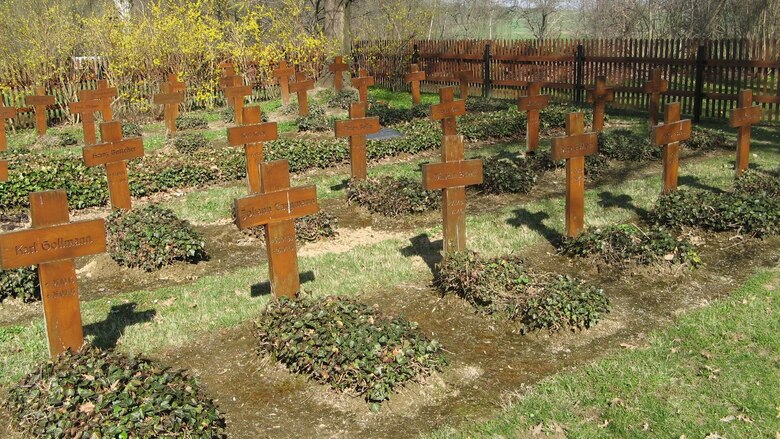 Das Bild zeigt einen kleinen, gepflegten Soldatenfriedhof mit mehreren Reihen einfacher Holzkreuze, die als Grabmale dienen. Jedes Grab ist mit einem Namensschild versehen, auf dem die Namen der Gefallenen zu lesen sind. Die Gräber sind von niedrigen Pfla