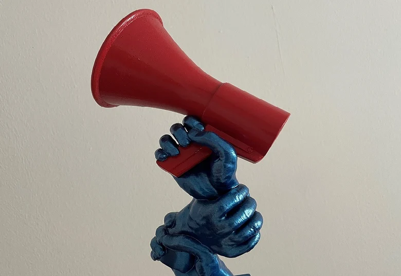Detailaufnahme einer Preisstatuette: Eine blaue, stilisierte Hand hält ein rotes Megafon nach oben vor hellem, neutralem Hintergrund.