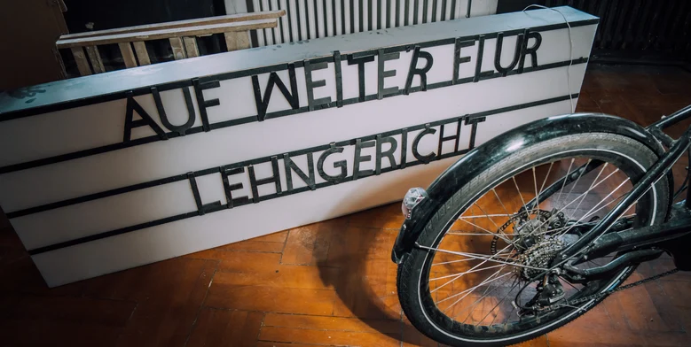 Das Bild wird durch ein Schild ausgefüllt. Auf diesem steht auf weiter Flur Lehngericht. Rechts unten ist ein Hinterrad eines Fahrrades abgebildet.