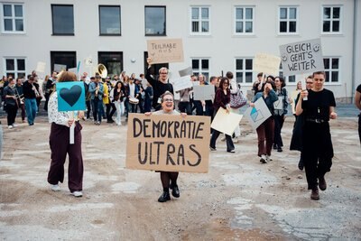 Auf dem Bild kommen Menschen auf den Betrachter zu. Sie haben Schilder in den Händen. Eine Frau steht im Bild-Mittelpunkt. Auf ihre Schild steht Demokratieultras.