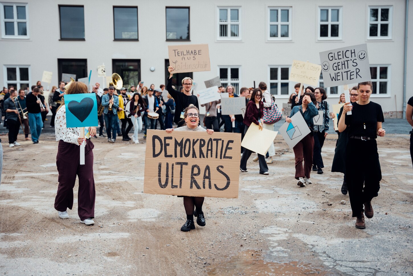 Auf dem Bild kommen Menschen auf den Betrachter zu. Sie haben Schilder in den Händen. Eine Frau steht im Bild-Mittelpunkt. Auf ihre Schild steht Demokratieultras.