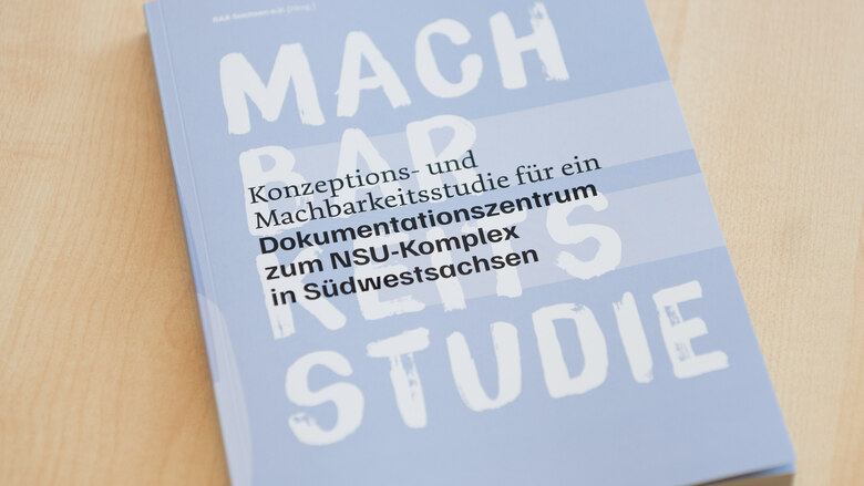 Auf einem Holztisch liegt eine hellblaue Broschüre mit dem Titel „Konzeptions- und Machbarkeitsstudie für ein Dokumentationszentrum zum NSU-Komplex in Südwestsachsen“, darüber groß der weiße Schriftzug „MACHBARKEITSSTUDIE“.