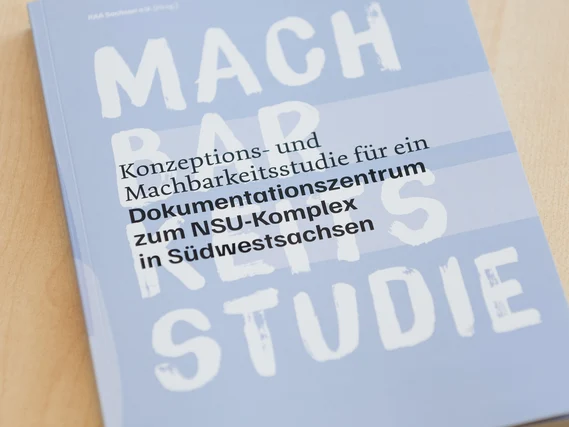 Auf einem Holztisch liegt eine hellblaue Broschüre mit dem Titel „Konzeptions- und Machbarkeitsstudie für ein Dokumentationszentrum zum NSU-Komplex in Südwestsachsen“, darüber groß der weiße Schriftzug „MACHBARKEITSSTUDIE“.
