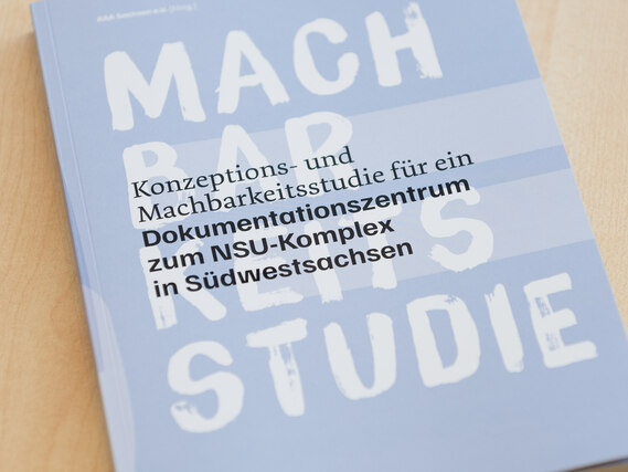 Auf einem Holztisch liegt eine hellblaue Broschüre mit dem Titel „Konzeptions- und Machbarkeitsstudie für ein Dokumentationszentrum zum NSU-Komplex in Südwestsachsen“, darüber groß der weiße Schriftzug „MACHBARKEITSSTUDIE“.