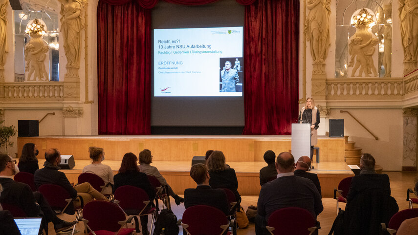 Im Hintergrund eine Leinwand mir dem Tirelmotiv der Veranstaltung. Am Pult eine Frau. Vor ihr sitzen mehrere Menschen mit dem Rücken zum Betrachter.
