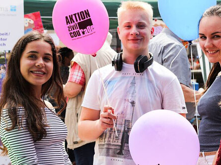 Drei Personen bei einer Veranstaltung mit pinken und hellblauen Luftballons mit Aufschrift „Aktion Zivilcourage".