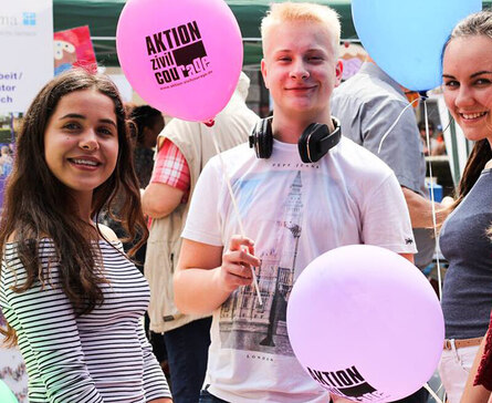 Auf dem Bild sind mehrere junge Menschen bei einer Outdoor-Veranstaltung zu sehen. Sie halten bunte Luftballons in den Händen, die mit dem Aufdruck „AKTION ZIVILCOURAGE“ versehen sind. Eine Person trägt ein weißes T-Shirt mit einem London-Motiv und hat Ko