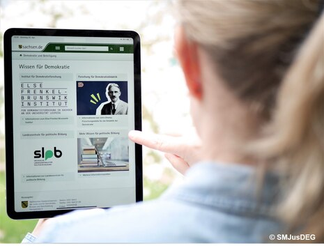 Das Bild zeigt eine Frau von hinten, die auf ein Tablet schaut. Auf diesem ist die Internetseite "Wissen für Demokratie" geöffnet.