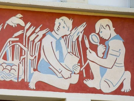 Wandbild im Stil der DDR: Zwei Kinder sitzen und knien zwischen Schilf, eines hält ein Buch, das andere untersucht Pflanzen mit einer Lupe, links ein Vogel über einem Nest; ausgeführt in Beige und Hellblau auf rotem Grund.